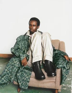 Model seated in an armchair wearing a patterned green coat and black leather boots for L'Officiel Malaysia Indoor Situation editorial