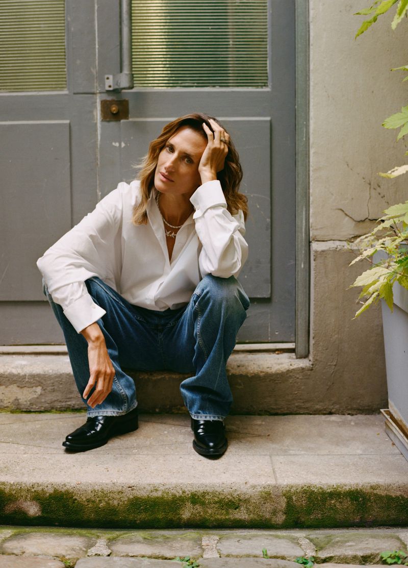 Woman with shoulder-length hair poses outdoors wearing a white shirt and blue jeans, sitting on concrete steps in a natural pose.