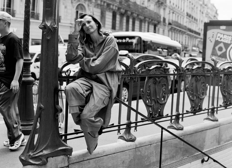 Woman in oversized clothing poses on a railing with a European city street, cars, buildings, and metro signage in the background.