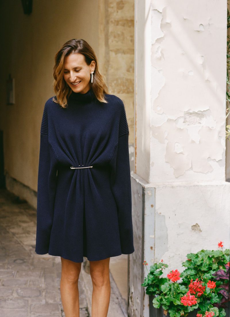 Adult woman in a dark blue sweater dress stands in an outdoor setting with red flowers on a partly cloudy day. She is smiling.