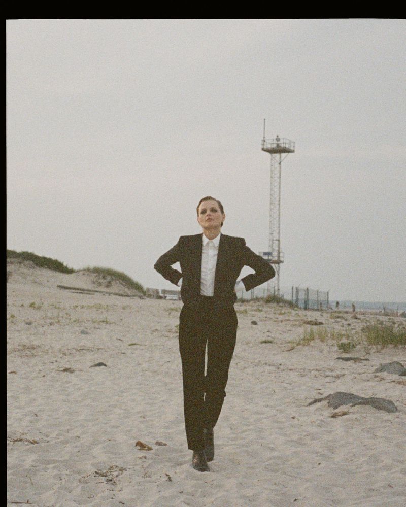 A woman in a black suit walks on a beach with a tower and dune grass in the background under an overcast sky, fashion portrait.