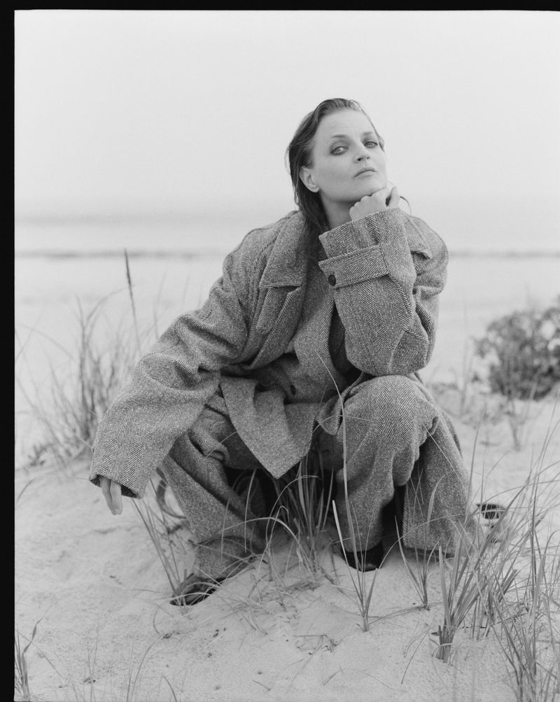Woman in tweed suit crouching on sand. Model poses on a beach with sea grass and an overcast sky in a black and white photograph.
