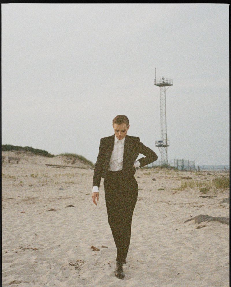 An adult female wearing a black suit and white shirt walks on a sandy beach under an overcast sky with a metal tower visible.