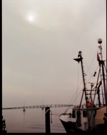 Blurred view of a fishing boat docked near a pier under an overcast sky, showing the boat's equipment and the pier in the background.