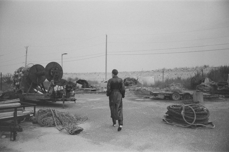 Monochrome shot of a person walking on a concrete surface surrounded by fishing gear and trailers, beneath an overcast sky.