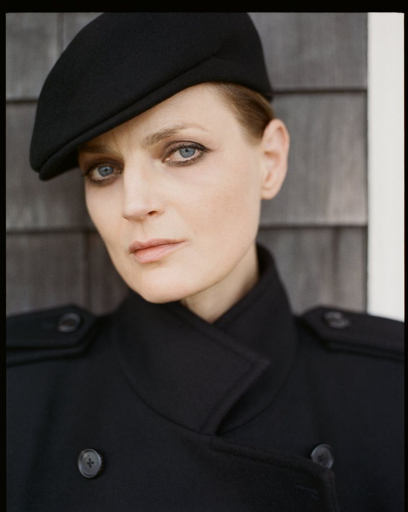 Model Guinevere van Seenus wearing a black cap and double-breasted coat against a wooden wall.