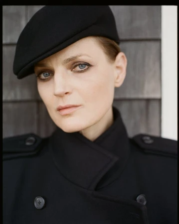 Model Guinevere van Seenus wearing a black cap and double-breasted coat against a wooden wall.