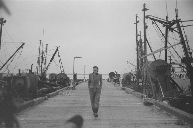 Monochrome photograph of an adult female in a suit walking on a dock flanked by docked fishing vessels on a cloudy day.