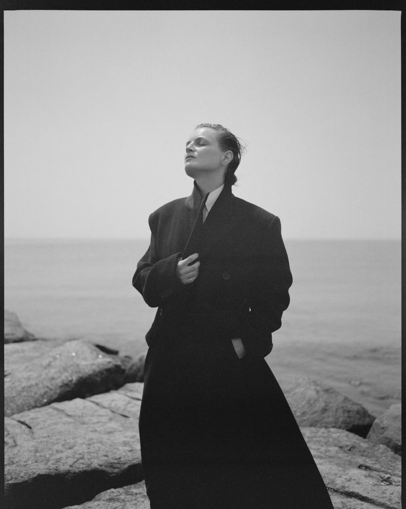 Monochrome portrait of a woman in a dark coat standing on rocks near the ocean, looking up, bathed in natural light.