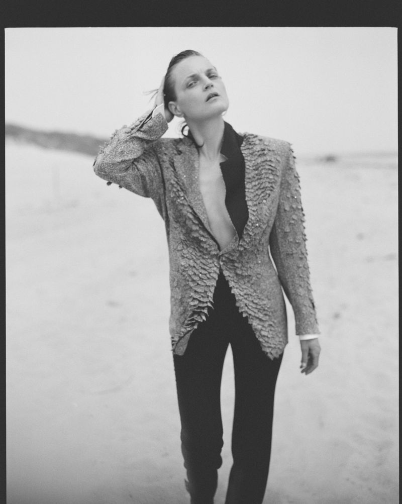 Guinevere van Seenus posing on a sandy beach wearing a textured blazer with structured shoulders and dark trousers in black and white.