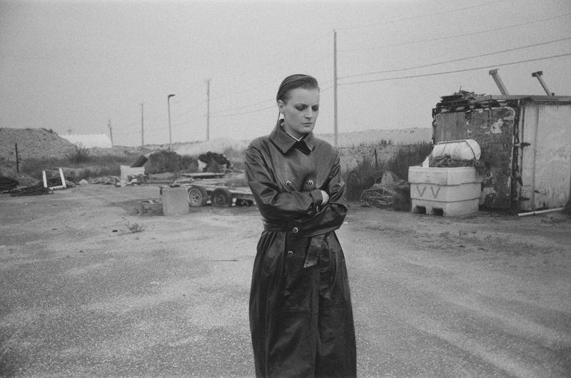 Black and white shot of a young adult in a belted trench coat standing on a dirt lot, with industrial structures and utility poles.