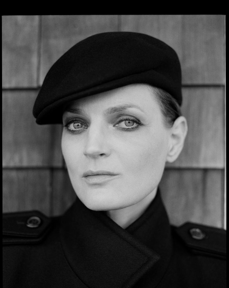 Monochrome portrait of a woman with fair skin wearing a black beret and coat, posing against a textured wood-paneled wall.