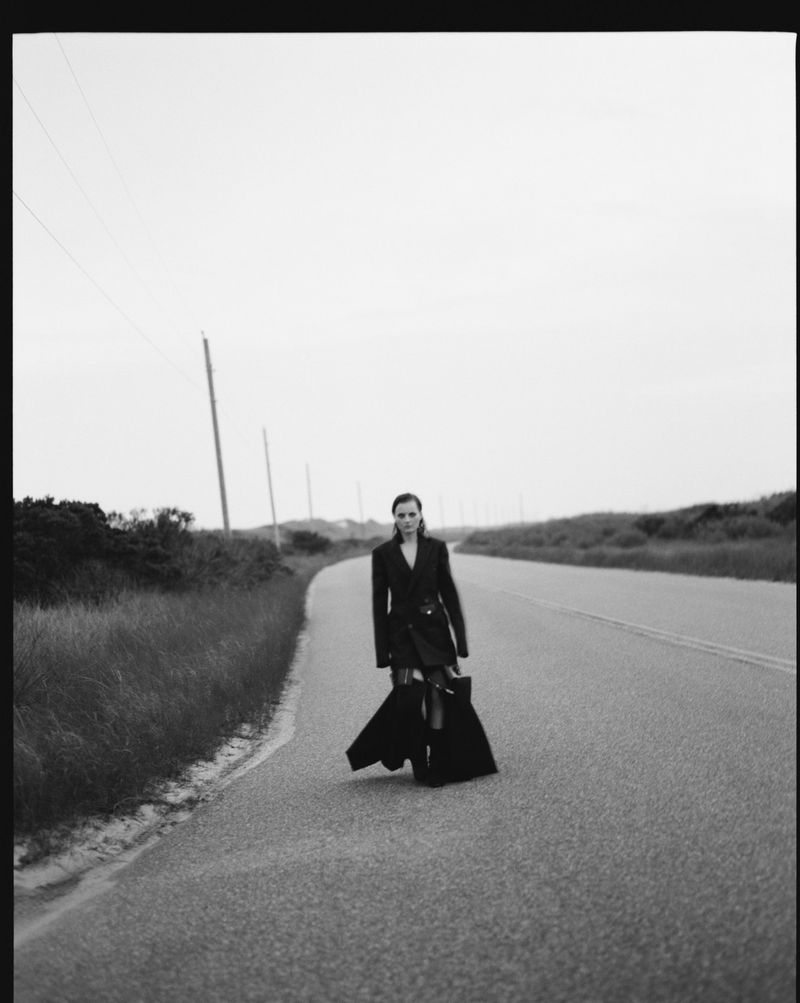Black and white fashion shot of a woman in a dark coat and skirt standing on a rural highway with vegetation in the background.