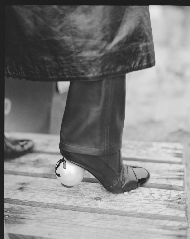 Woman wearing black leather boots with a round heel, standing on wooden planks, and wearing a shiny leather coat.