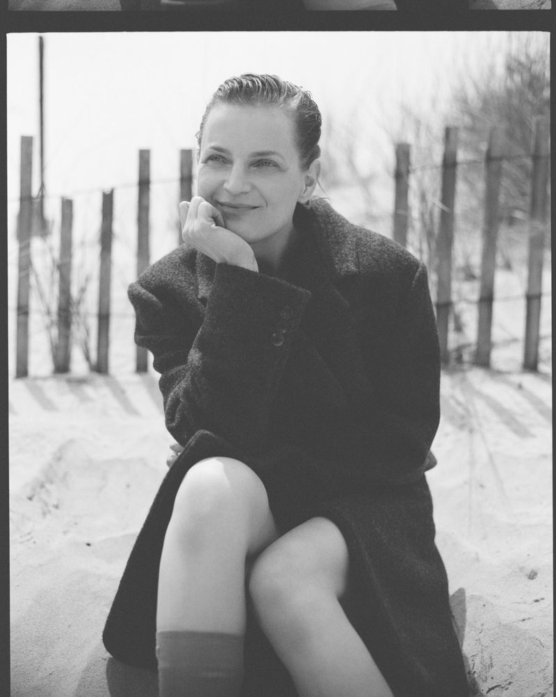Black and white portrait of a woman sitting on sand wearing a coat, looking off into the distance next to a wooden fence.
