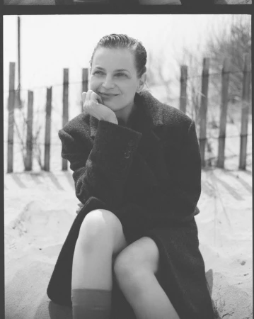Black and white portrait of a woman sitting on sand wearing a coat, looking off into the distance next to a wooden fence.