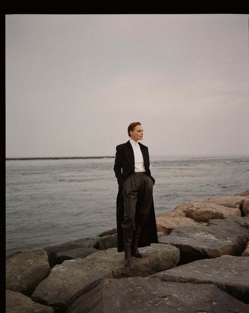 Woman in a black coat and grey trousers stands on a rock breakwater by the ocean with a grey sky. Fashion editorial photography.