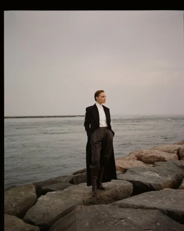 Woman in a black coat and grey trousers stands on a rock breakwater by the ocean with a grey sky. Fashion editorial photography.