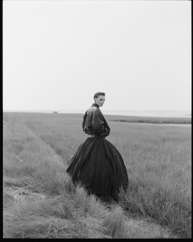 Woman in black dress standing in a grassy field under overcast sky. The landscape is monochromatic in this outdoor fashion photograph.