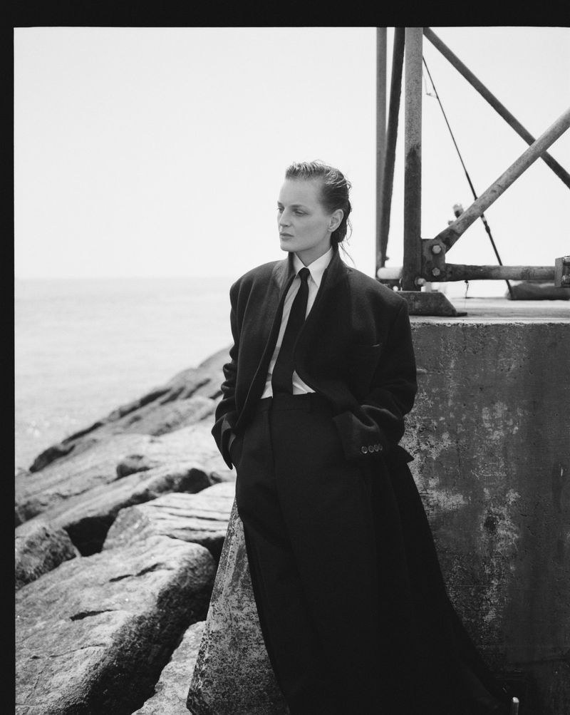 Black and white portrait of a woman in a suit and tie, leaning against a stone structure by the sea, gazing away with a neutral expression.