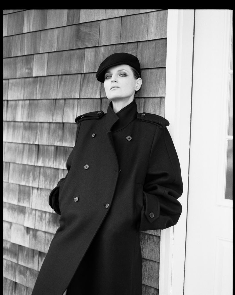 Monochrome portrait of a female adult wearing a coat and hat leaning against a wall with wood cladding and a white door frame.