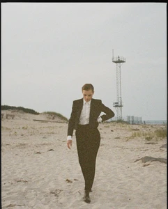 Model Guinevere van Seenus wearing a black suit and white shirt walking on a sandy beach under an overcast sky.