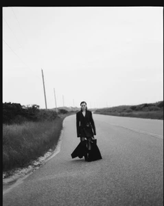 Guinevere van Seenus in a black tailored coat standing on an open asphalt road lined with coastal dunes in black and white.