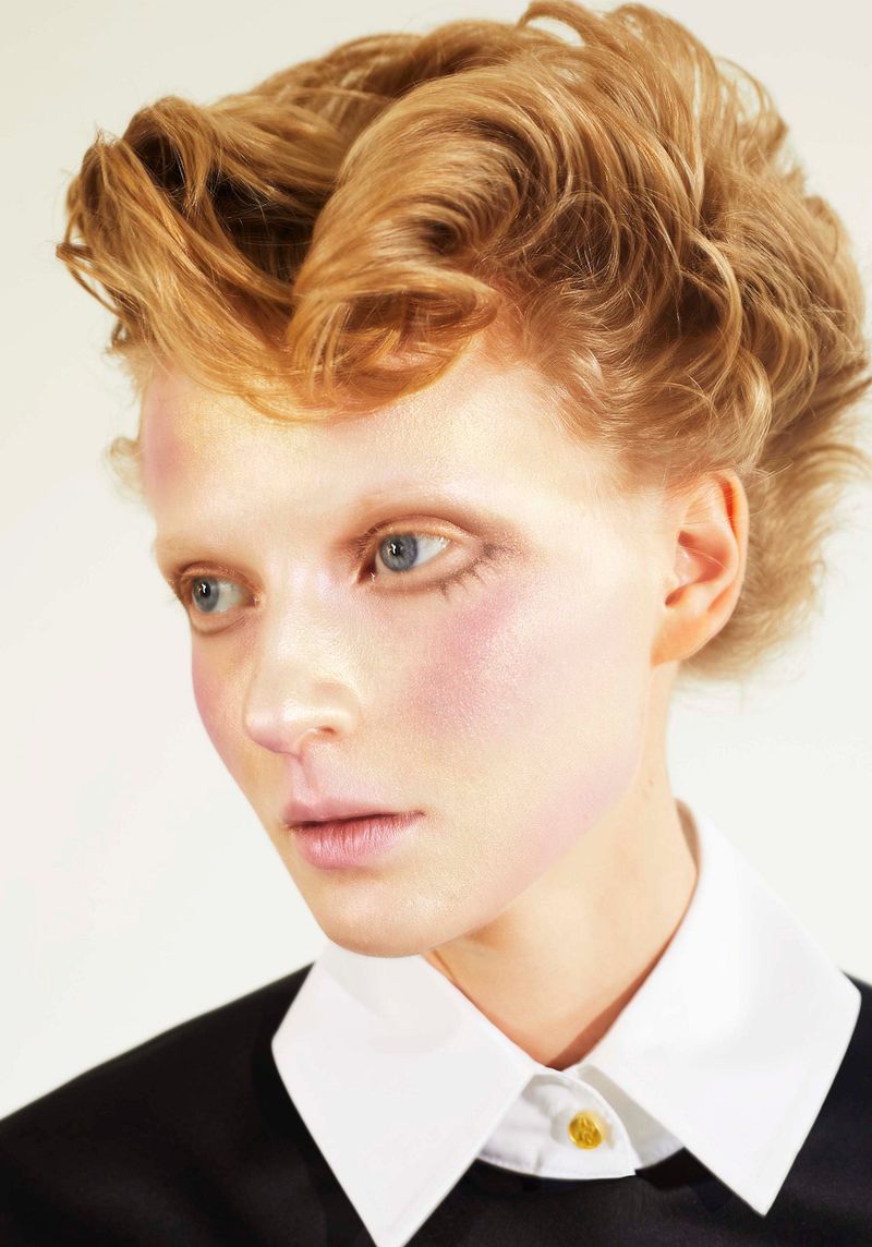 Portrait of a blonde woman with styled hair wearing a black shirt with a white collar against a light background in a beauty shot.