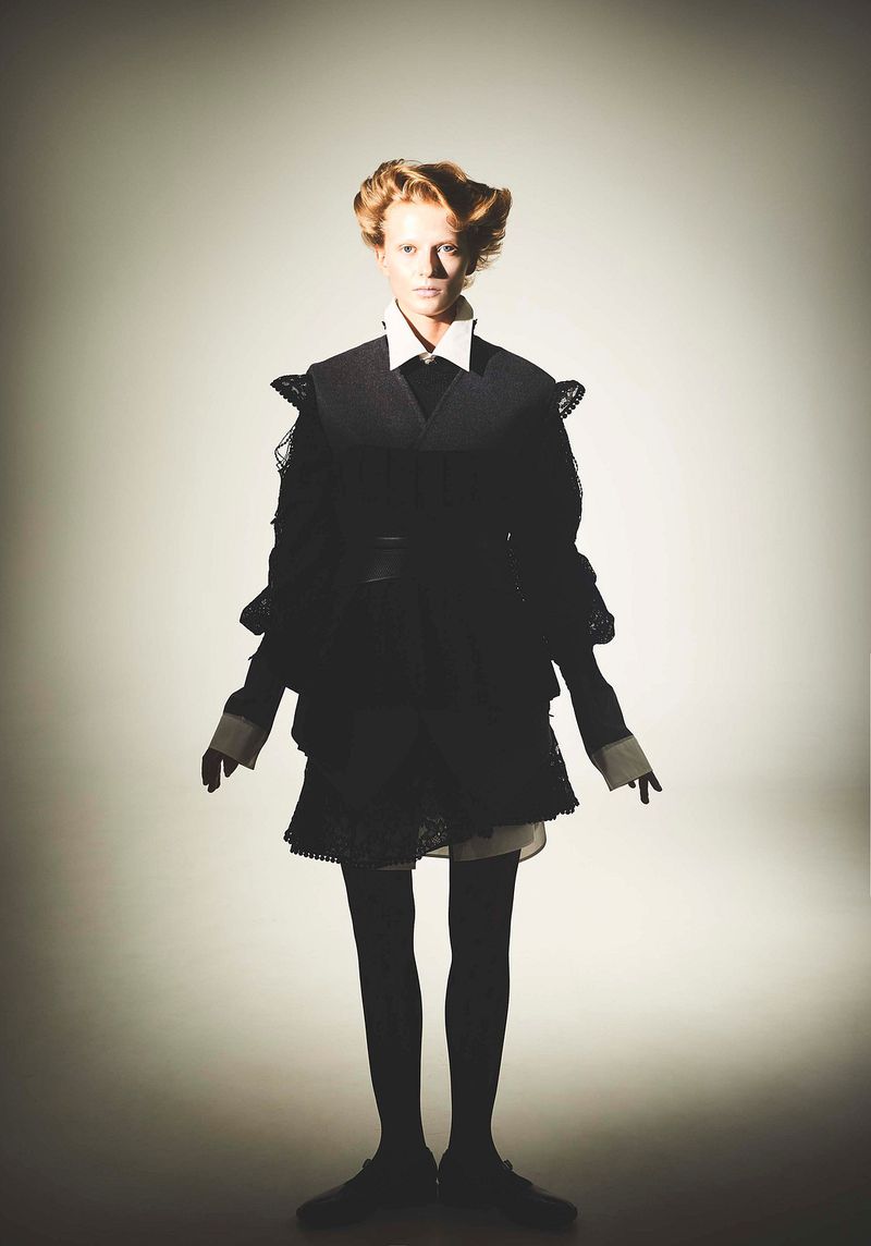 A model stands in a photo studio wearing a black two-piece garment and black tights against a bright neutral background in a full shot.