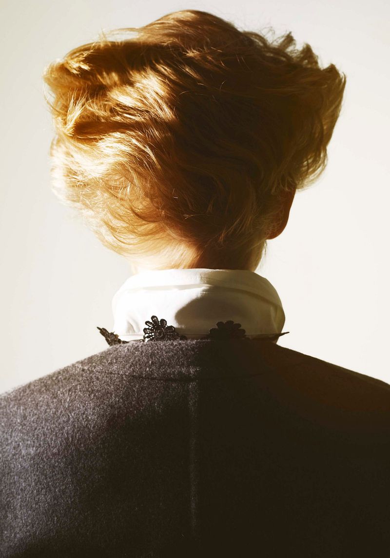 Blonde hair fashion detail with a gray jacket and white collared shirt, viewed from behind in a brightly lit studio setting.