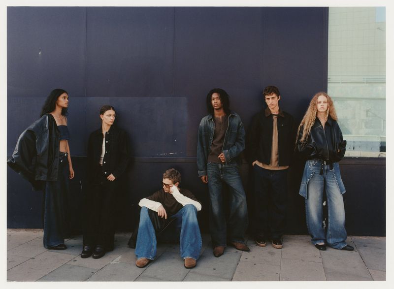 Six people stand in a line in front of a dark blue wall, wearing fashionable clothing; one person is sitting on the ground.