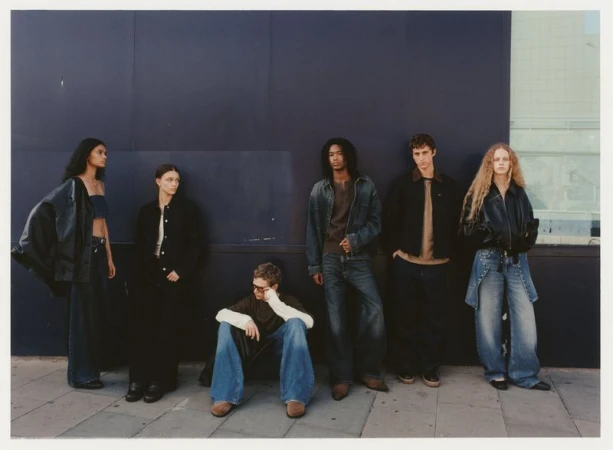 Six people stand in a line in front of a dark blue wall, wearing fashionable clothing; one person is sitting on the ground.