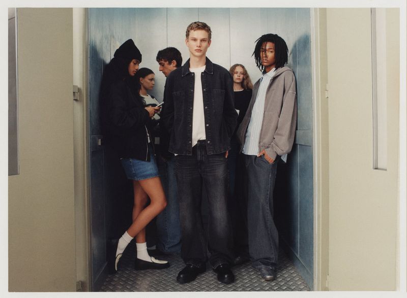 Six young adults pose in an elevator, showcasing casual fashion with denim, hoodies, and sneakers, set against a neutral-toned interior.