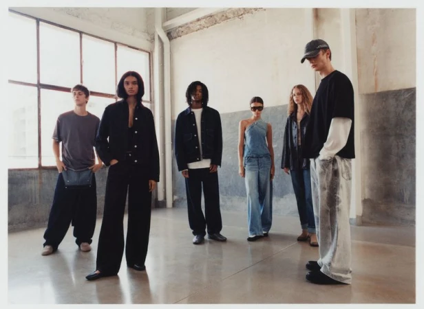 Six people stand in a semi-circle in a large, open room with neutral tones, wearing casual clothing and posing for a fashion shoot.