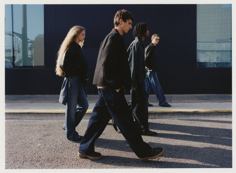 A group of four people walking on a sidewalk in an urban environment. They are wearing casual clothing and walking in the same direction.