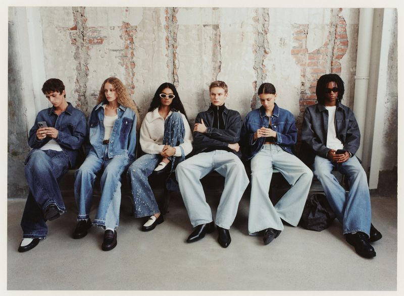 Six young adult fashion models sit on a bench against a distressed wall, all wearing denim outfits in a studio setting.