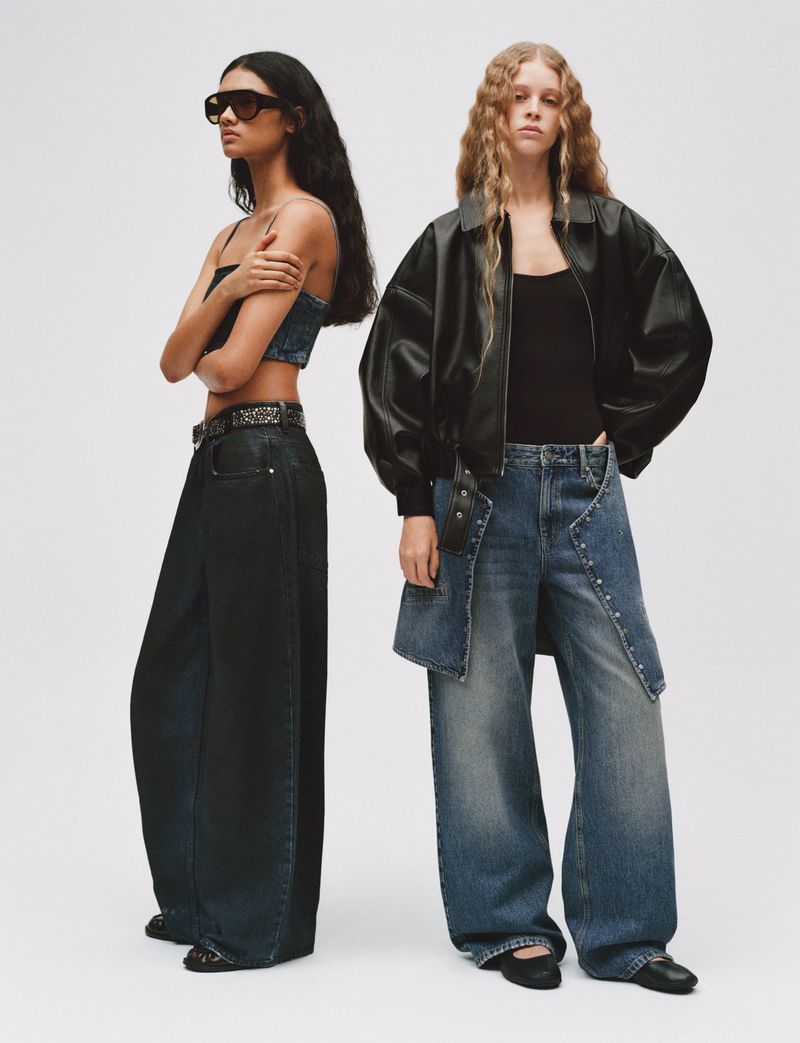 Two female fashion models pose in wide-leg jeans and tops in a studio, one with sunglasses, under bright lighting and neutral background.