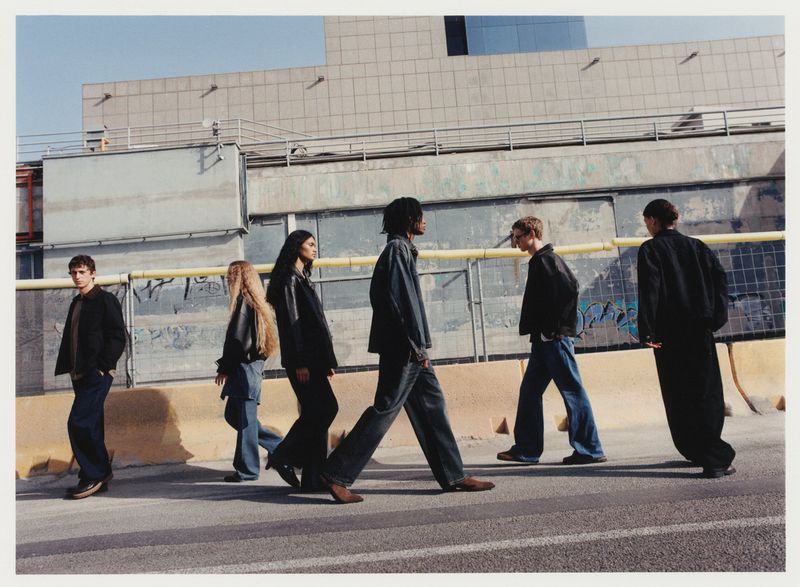 Six people walk across a street in casual wear, with an urban backdrop of a large, light-colored commercial building under a blue sky.