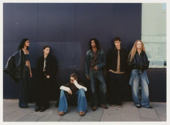 Six models in denim and dark jackets posing along an outdoor dark wall on a paved sidewalk for Pull & Bear.