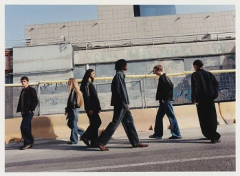 Group of models walking along an asphalt street outdoors in front of a concrete building exterior and yellow barrier.