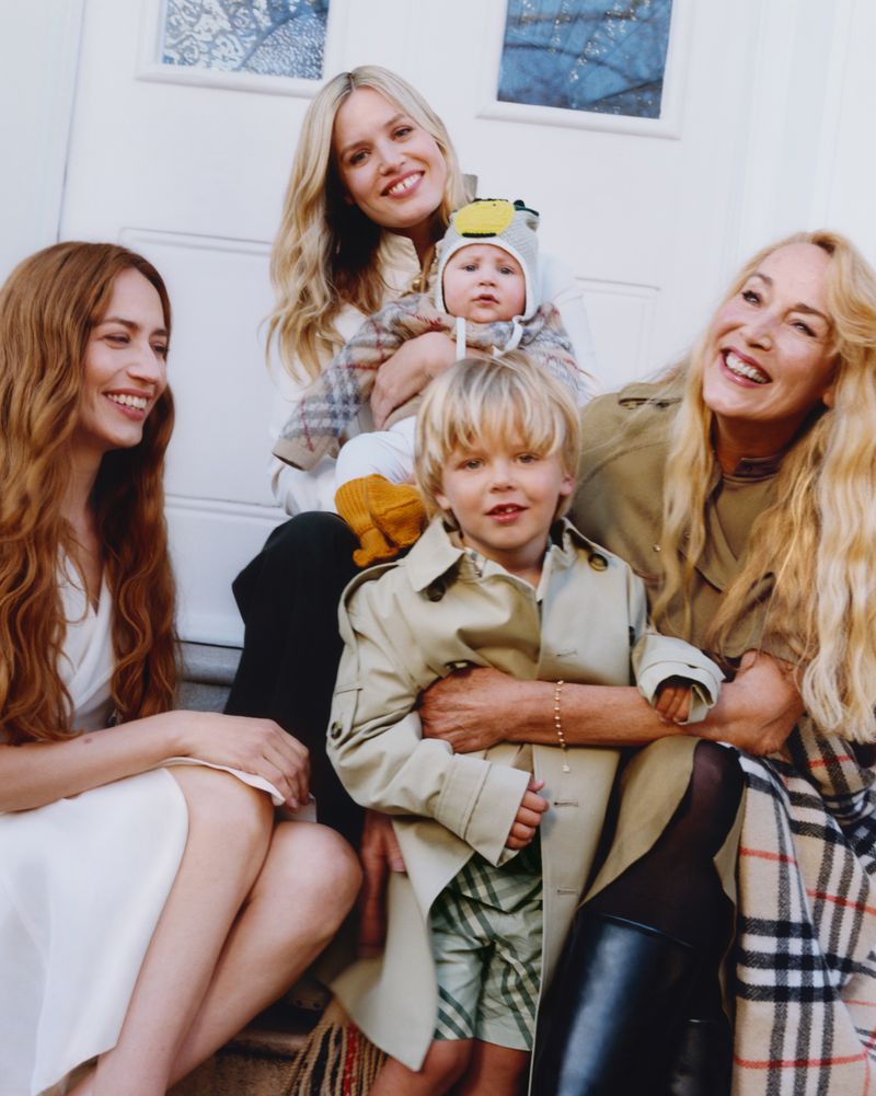 Smiling family group portrait of three women and two children in stylish trench coats and beige plaid fabrics in an indoor setting.