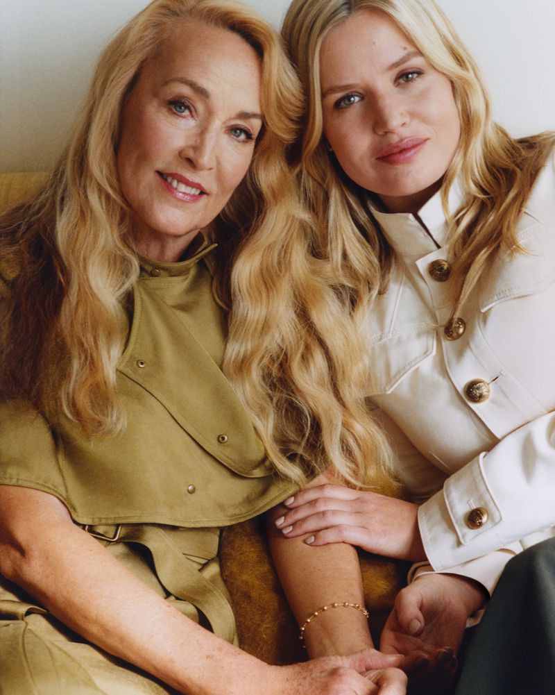 Two women, one middle-aged and one younger, are posing indoors. The women have long blond hair and are wearing elegant neutral-toned clothing.