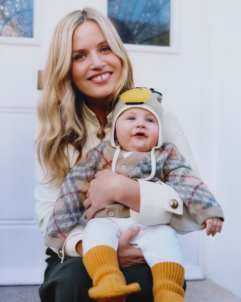 A smiling woman with long blonde hair holds a baby with a knitted hat and mustard-yellow socks, sitting on a white porch.