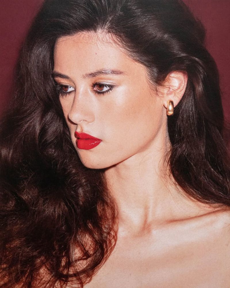 Portrait of a woman with dark wavy hair, red lipstick, and gold hoop earrings against a reddish-brown backdrop in an indoor setting.