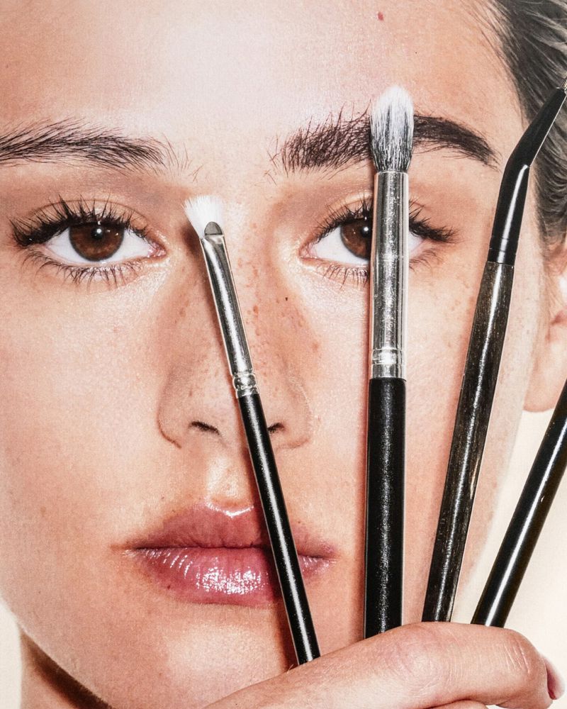 Close-up portrait of a woman's face surrounded by makeup brushes, focusing on eyes, skin texture, and cosmetic tools.