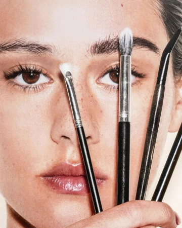 Close-up portrait of a woman's face surrounded by makeup brushes, focusing on eyes, skin texture, and cosmetic tools.