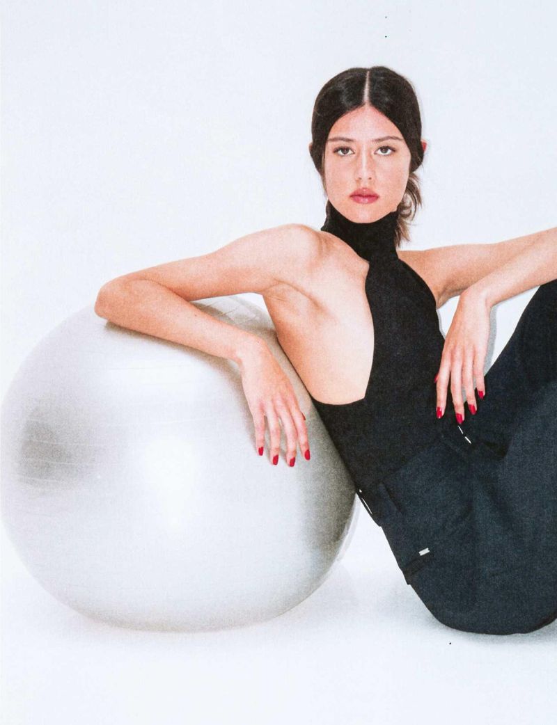 Young woman in black top and dark pants poses next to silver exercise ball in a brightly lit white studio setting for a fashion shoot.