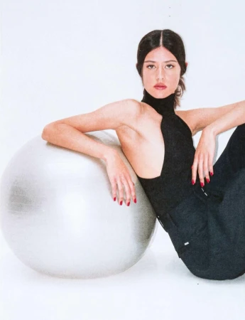 Young woman in black top and dark pants poses next to silver exercise ball in a brightly lit white studio setting for a fashion shoot.