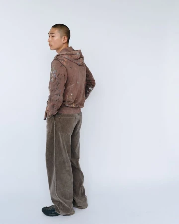 Young adult male in hooded sweatshirt and wide-leg pants standing in a photo studio, looking over his shoulder.