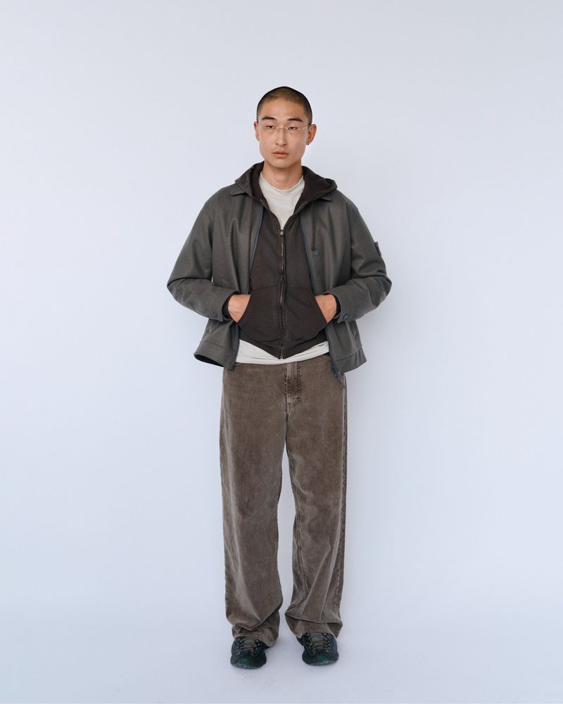 Full-length studio shot of a young adult male in a brown jacket, vest, and corduroy pants against a plain white backdrop.
